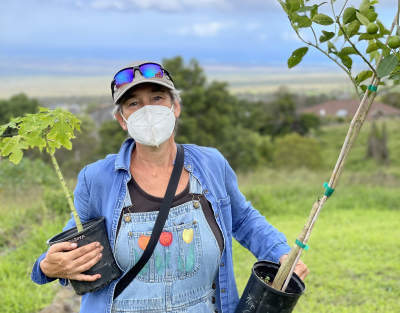 A woman in a mask and overalls holds two saplings.