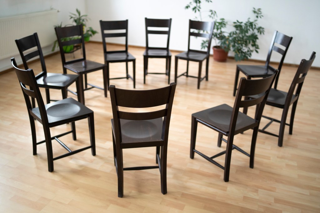 A circle of empty chairs in a plain room with a wooden floor