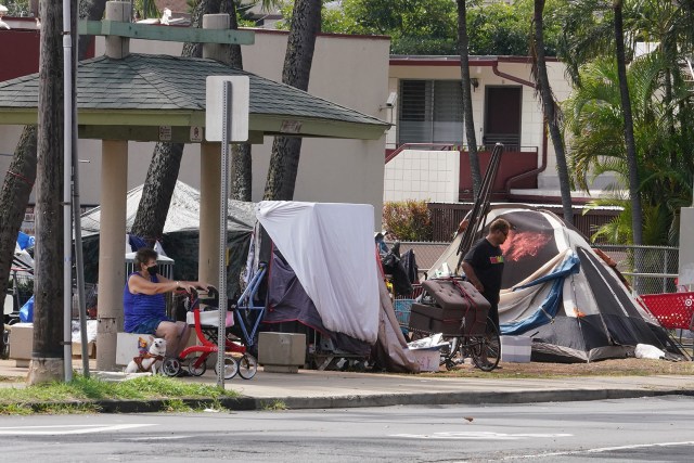 TheBus customer sits at the bus stop on Kalakaua Avenue near South King Street with a nearby homeless encampment.