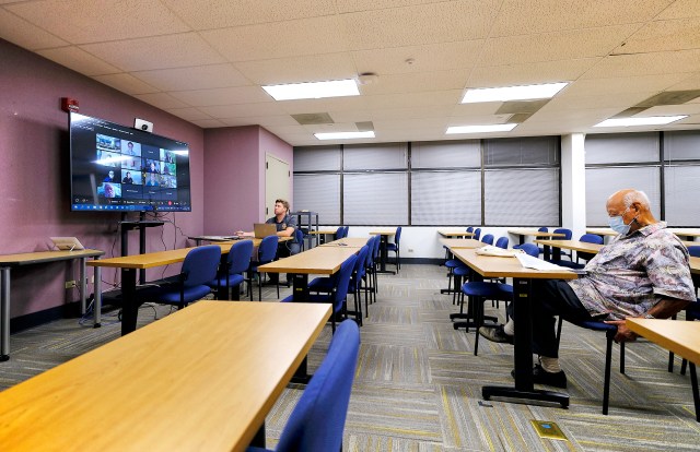 Desks face a large TV screen of Webex participants. One person sits there, studying the papers in front of him. In the background, a person controls the computer.