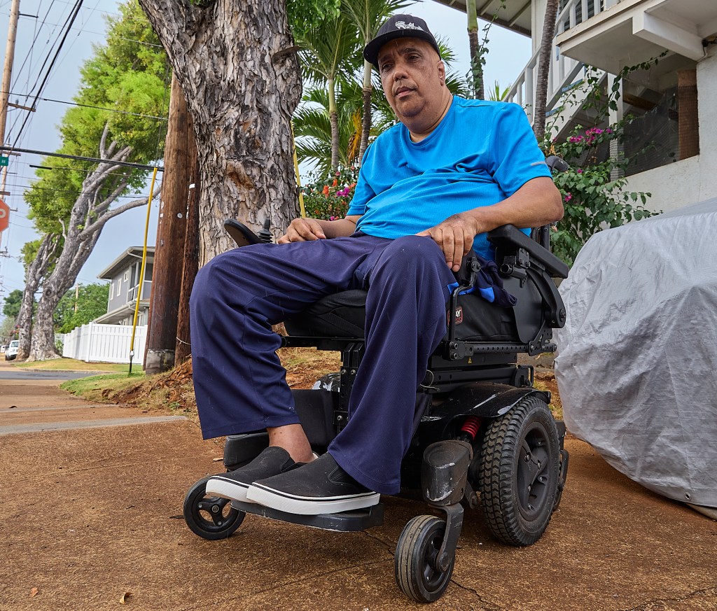 Ryan Smith sits outside his home next to the tree whose roots continue to force the sidewalk upward and impede his ability to maneuver his wheelchair