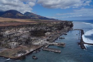 The ruins of Lahania town eerily rests calmly as a large wave breaks over Lahaina Harbor breakwall Thursday, Aug. 10, 2023, in Maui. Two days prior, a large, fast-moving wildfire consumed this historical West Maui town. (Kevin Fujii/Civil Beat/2023)