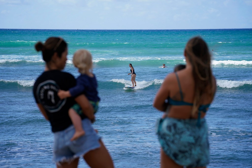Surfing Mom’s Sachiko Stokes, from left, holds Daniel Ritchie, while his mom Kelli Ritchie surfs and Santiago Martin watches in the arms of Victoria Moody during the group’s weekly outing Tuesday, Aug. 15, 2023, at White Plains Beach in Ewa Beach. The moms take turn surfing while other moms watch the keiki. (Kevin Fujii/Civil Beat/2023)