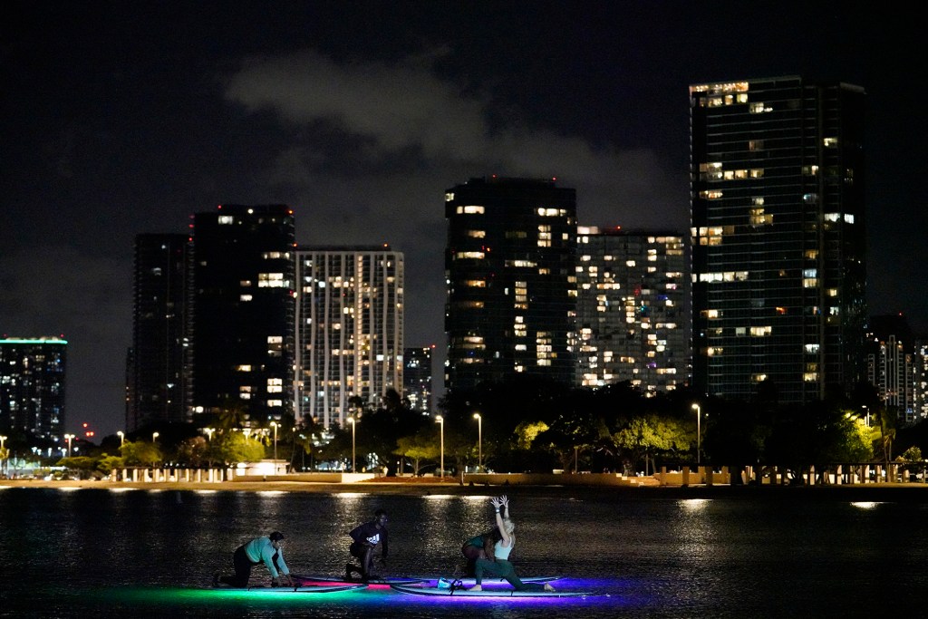 Honeymooners Prashina Siva, from left, her husband Kumar Facey, both from Toronto, Teondra Mills of Honolulu and Yoga Floats instructor Julie Garvilla practices a Fireworks Glow and Flow yoga session Friday, Nov. 17, 2023, at Ala Moana Beach Park in Honolulu. Lights under the SUPs create a glow under their boards. After their practice, they watched the Friday night fireworks over Waikiki. (Kevin Fujii/Civil Beat/2023)