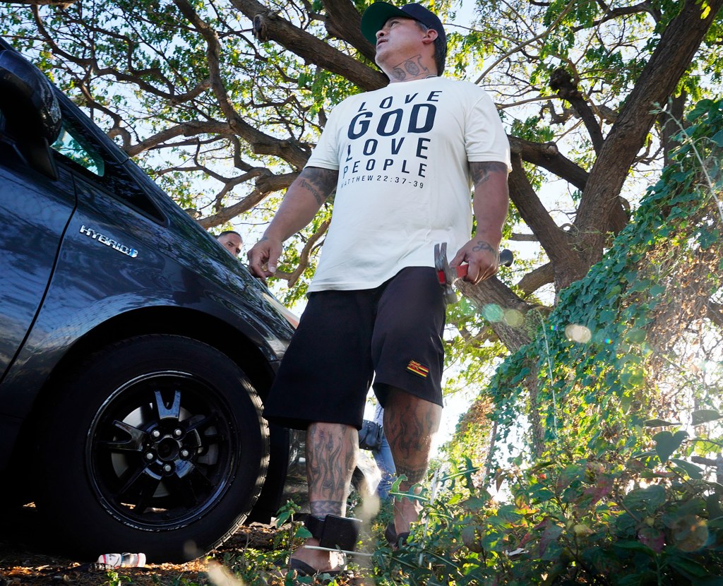 Mana Olayan stands and turns to his bible study group, “God Forgives Bad Boys and Bad Girls,” after pounding a stake into a banyan tree’s root Tuesday, June 6, 2023, in Ewa Beach, Hawaii. Olayan said this tree was the focal point of the neighborhood’s low point. Gang members hung out here, sold drugs and guns and created trouble according to Olayan.