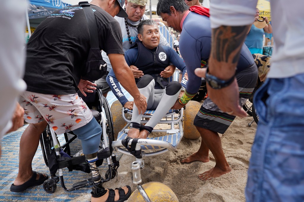 Volunteers and fellow competitors move Tomoki Fujiwara of Japan onto an all-terrain wheelchair for his 2024 Hawaii Adaptive Surfing Championships Prone Assist division heat Monday, May 13, 2024, in Honolulu. (Kevin Fujii/Civil Beat/2024)