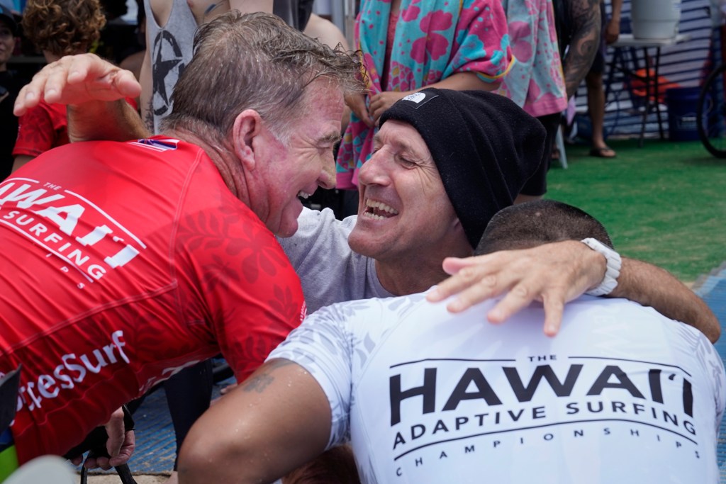 Any Knee Kneeling men competitors Mark “Mono” Stewart, wearing red rash guard, and Masafumi Kobayashi, wearing white rash guard, get a hug from Wave Ski surfer Spike Kane after their 2024 Hawaii Adaptive Surfing Championships heat takes place Tuesday, May 13, 2024, in Honolulu. The three competitors shared a house at the first Association of Adaptive Surfing Professionals competition of the year in Australia this past March. (Kevin Fujii/Civil Beat/2024)