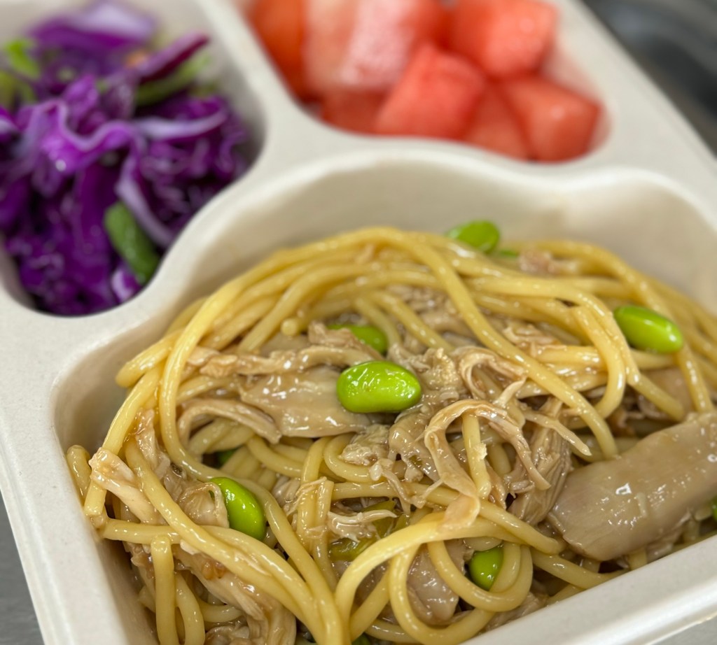 A school meal from Happy Bento consists of chicken sesame noodles and fresh fruits and vegetables.