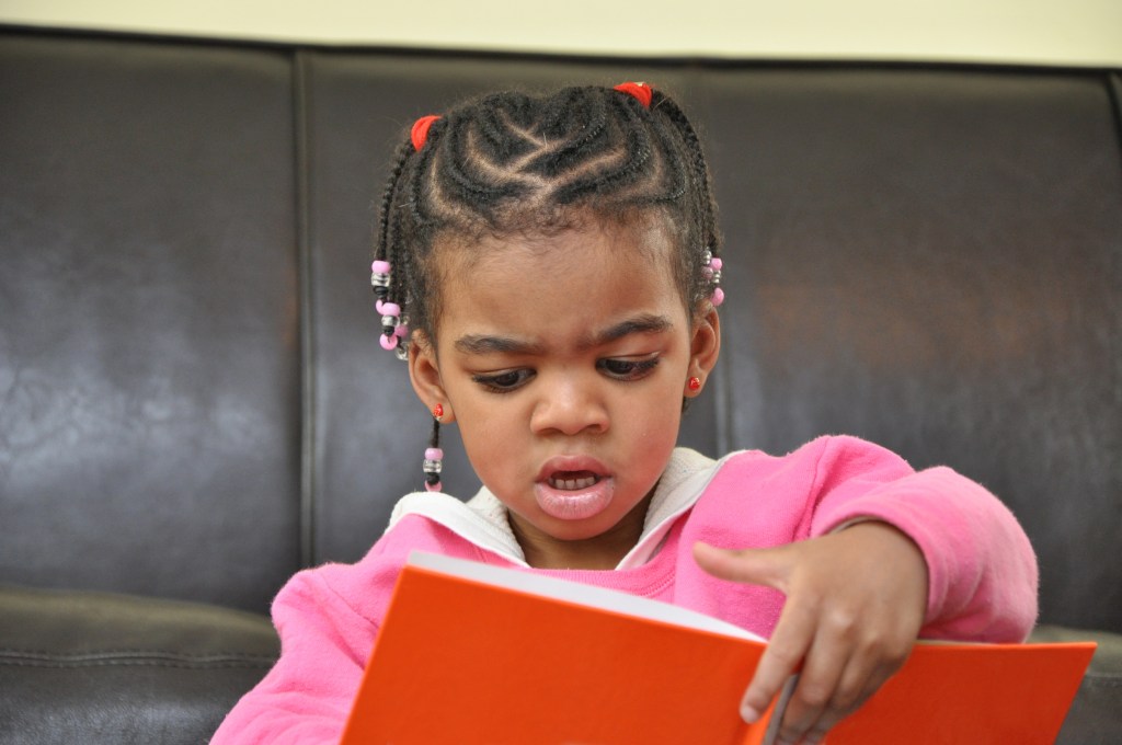 Little Girl with corn row hair style looking down making face reading a book