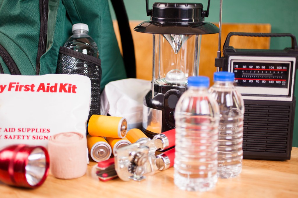 Emergency preparedness supplies. A large pile of supplies to be used in case of a natural disaster (hurricane, flood, earthquake, etc.) including: flashlight, backpack, batteries, water bottles, first aid kit, lantern, radio, can opener, mask.  No people.