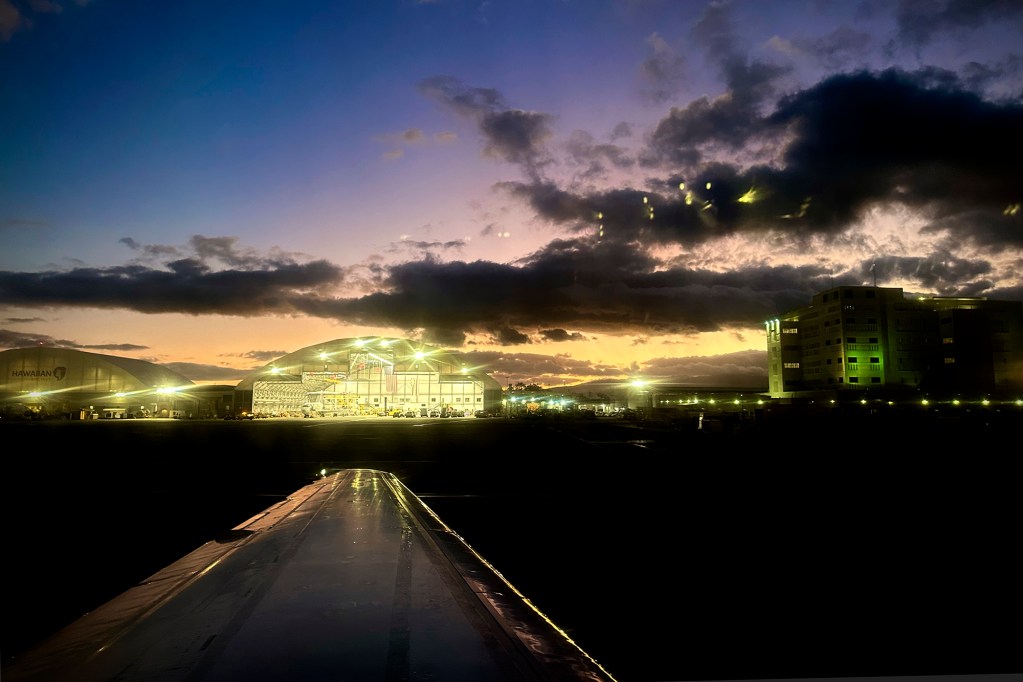 A Hawaiian Airlines Boeing 717 port wing points to their hanger as the sun sets Friday, June 28, 2024, at Daniel K. Inoyue International Airport in Honolulu. Reflections from inside the airplane appear to hover in the clouds. (Kevin Fujii/Civil Beat/2024)