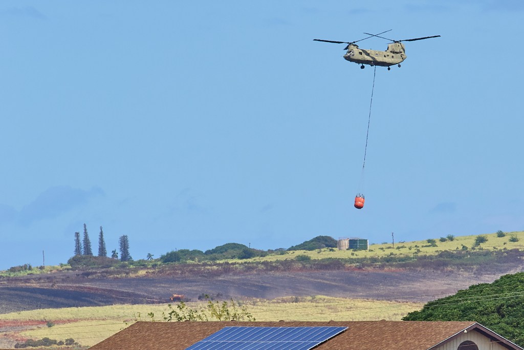 A Chinook helicopter works to douse hotspots just west of Moi Road in Hanapepe, Kauai. The fire was considered contained by daybreak on July 16th but crews spent the overnight hours checking for hot spots and protecting the community from flare-ups (David Croxford/CivilBeat/2024)