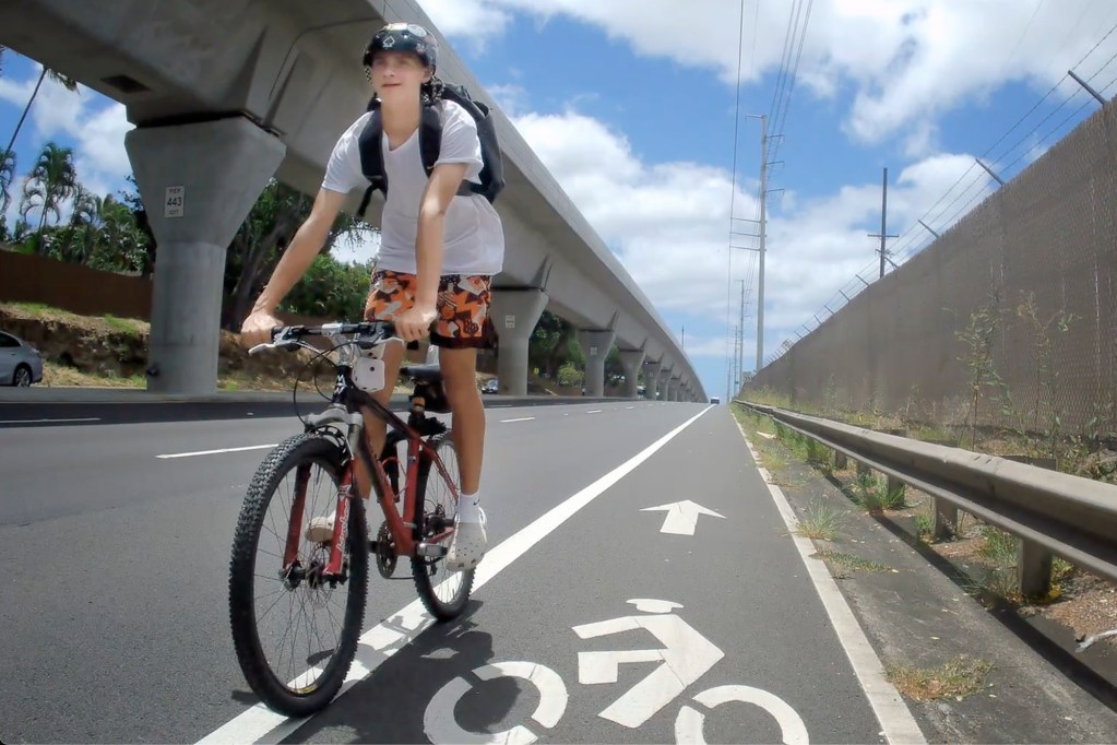 The sign couldn’t be clearer as a person rides a bike in the wrong direction on Kamehameha Highway near Pearl Harbor Saturday, Aug. 17, 2024, in Honolulu. His helmet isn’t buckled securely either. When his body hits the ground, the helmet will fly off, offering no protection. Still photo from video. (Kevin Fujii/Civil Beat/2024)
