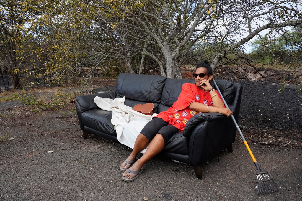 Hokuao Kaohelaulii sits on the sofa he slept on overnight after the previous day’s vegetation fire Monday, Sept. 16, 2024, in the Kalaeloa area of Ewa Beach while Honolulu Fire Department firefighters continue to monitor hot spots. Kaohelaulii, of Niihau, said his belongings were destroyed. He doesn’t call himself homeless as his home is on Niihau. Kaohelaulii is on Oahu to attend school and work. He said many homeless live in the area near Kalaeloa Airport. (Kevin Fujii/Civil Beat/2024)