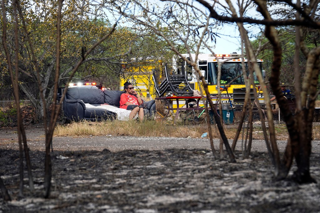 Hokuao Kaohelaulii sits on the sofa he slept on overnight after the previous day’s vegetation fire Monday, Sept. 16, 2024, in the Kalaeloa area of Ewa Beach while Honolulu Fire Department firefighters continue to monitor hot spots. Kaohelaulii, of Niihau, said his belongings were destroyed. He doesn’t call himself homeless as his home is on Niihau. Kaohelaulii is on Oahu to attend school and work. He said many homeless live in the area near Kalaeloa Airport. (Kevin Fujii/Civil Beat/2024)