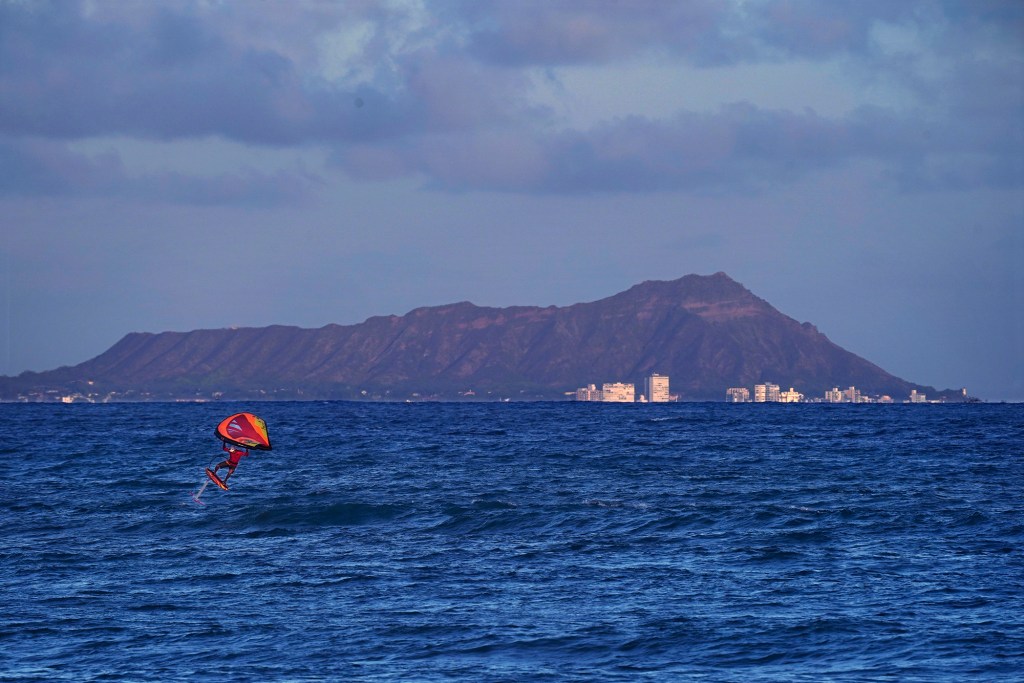 A wingfoiler catches air at White Plains Beach with Diamond Head rising above the horizon Thursday, Sept. 19, 2024, in Ewa Beach. The wing acts like a sail or kite to propel the athlete. The hydrofoil under the board lifts the board above the water when the rider reaches a certain speed. (Kevin Fujii/Civil Beat/2024)