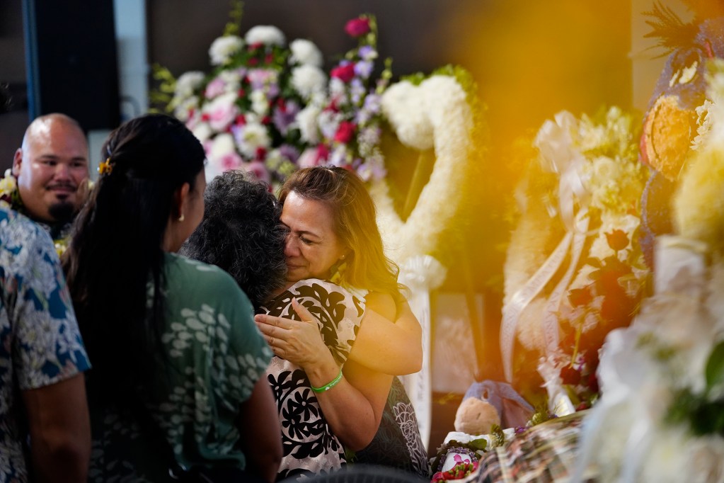 Courtney Raymond-Arakaki’s mother Chermaine Raymond gives a hug while boyfriend Wyman Keamo, left, looks on after her memorial service Sunday, Sept. 22, 2024, in Honolulu. The 34-year-old was killed in an ongoing dispute between neighbors in Waianae. A flower creates the orange glow on the right side of this photograph. (Kevin Fujii/Civil Beat/2024)