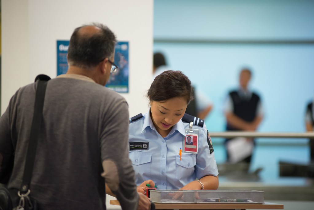 A woman in a blue uniform holds a marker in her hand, looking at a form while a man waits next to her.