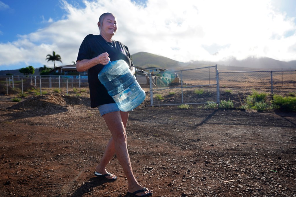 Susie Richter labors carrying a 5-gallon jug of water for the plants at the Lahaina memorial Wednesday, Oct. 2, 2024, in Lahaina. Water tanks were located closer to the memorial but has since been removed after Maui County declared public utilities water potable. (Kevin Fujii/Civil Beat/2024)