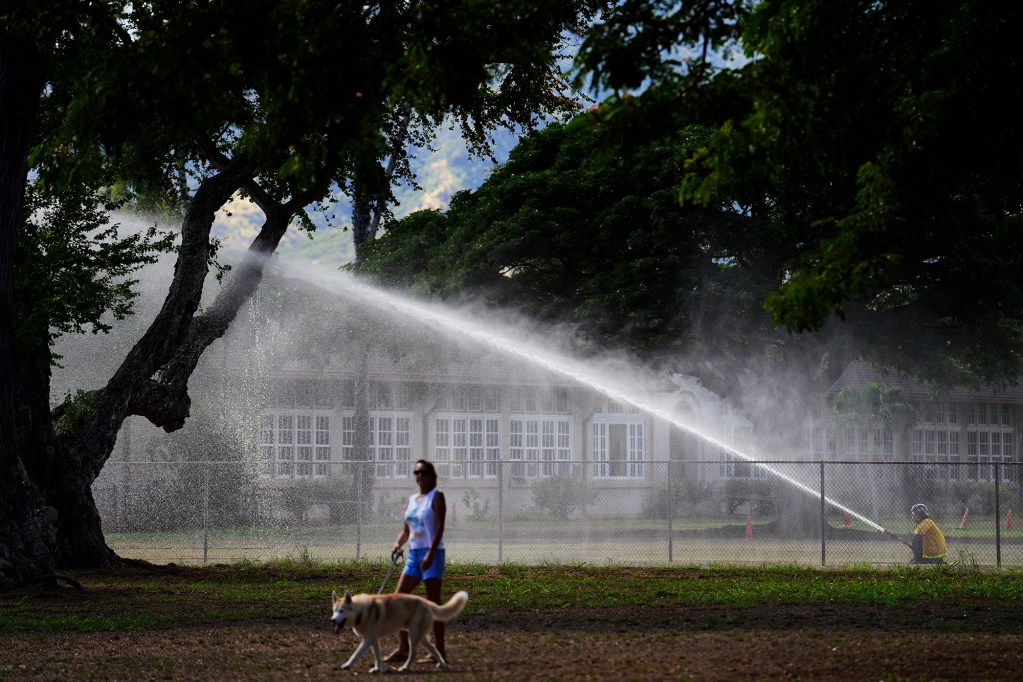 Kris Roberts and her rescued, almost 3-years-old, Siberian husky Dave stroll through Kaiaka Bay Beach Park as Honolulu Fire Department’s Waialua Engine 14 sprays water Monday, Oct. 7, 2024, in Haleiwa. Releasing the water fulfilled two purposes: the fire engine’s water tanks needed draining and rookies learned how to operate the pumps. (Kevin Fujii/Civil Beat/2024)