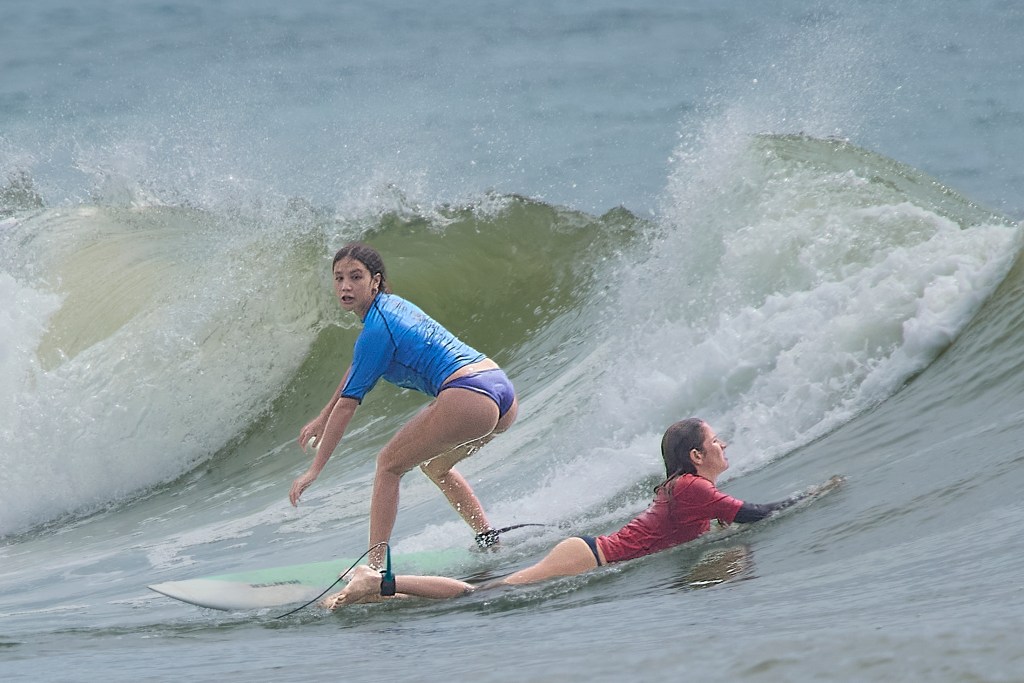 High School surfing started its season at Ala Moana Bowls on Saturday October 26th, 2024 with 17 schools competing in the day long event. In the girls short board event Kara Watson (Blue) takes off on a wave as Solbee Ahina (Red) paddles out.(David Croxford/Civil Beat/2024)