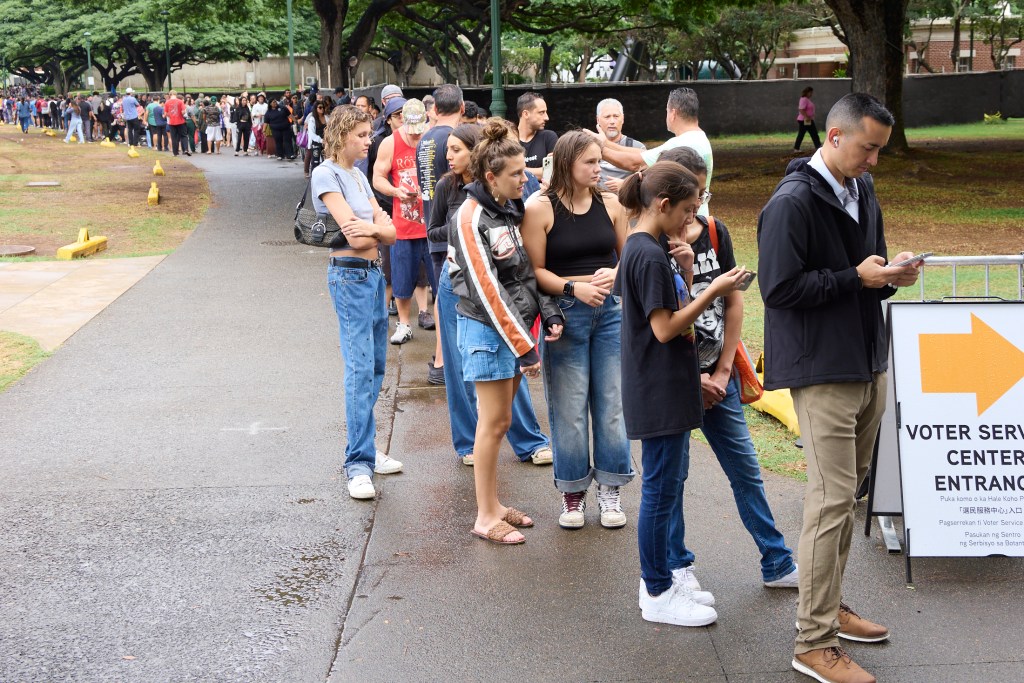 Voters at Honolulu Hale on King Street in Honolulu were met with Poll workers as they drove through the election only lane that had been set aside and coned to allow drivers to either hand off their ballots to Poll workers or drop them into several Ballot boxes placed and manned along King Street.  The lines snaked around the rear of Honolulu Hale, out to the walkway in the rear and then went all the way to Ala Wai and back providing wait times in excess of 2 hours (David Croxford/Civil Beat/2024)