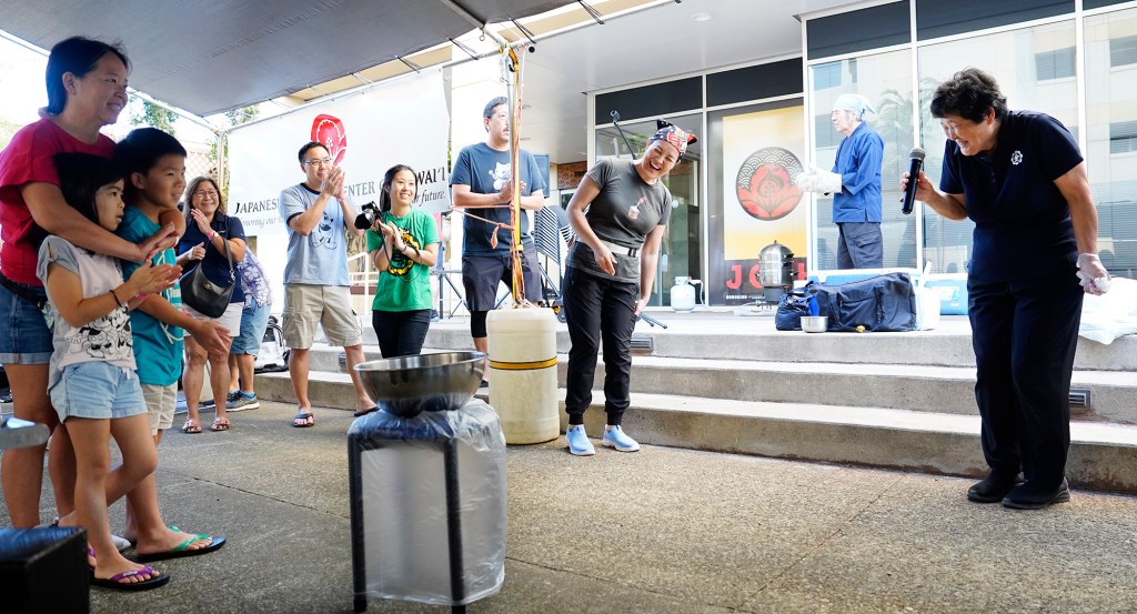 The Konko Mission of Wahiawa Rev. Reiko Yano, right, giggles with her daughter, Rev. Edna Matsuoka, third right, after singing a song traditionally sung before mochitsuke (mochi pounding) Saturday, Dec. 14, 2024, at the Japanese Cultural Center of Hawai’i in Honolulu. Head Rev. Yasuhiro Yano, second right, applauds with the crowd gathered for the mochitsuki demonstration. Mochitsuki is a traditional Japanese festival which gathers family and friends to celebrate the incoming year. Mochi is a celebrated food consumed for good luck, prosperity, unity, trust and good health for the new year. The Konko Mission of Wahiawa has been demonstrating mochitsuki since 1992. (Kevin Fujii/Civil Beat/2024)