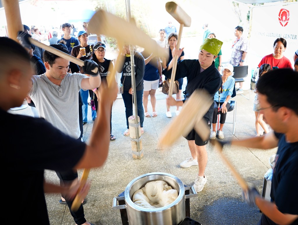 Konko Mission of Wahiawa volunteers blur with activity as they demonstrate a mochitsuke (mochi pounding) by pounding the mochigome (glutinous rice) with kine (wooden mallets) inside head Rev. Yasuhiro Yano’s custom-made usu (mortar) Saturday, Dec. 14, 2024, at the Japanese Cultural Center of Hawai’i in Honolulu. They are Evan Davison, from left, of Honolulu, Kirby Shiraishi of Ewa Beach, Nick Furusho and Clayton Matsuoka, both from Wahiawa. (Kevin Fujii/Civil Beat/2024)