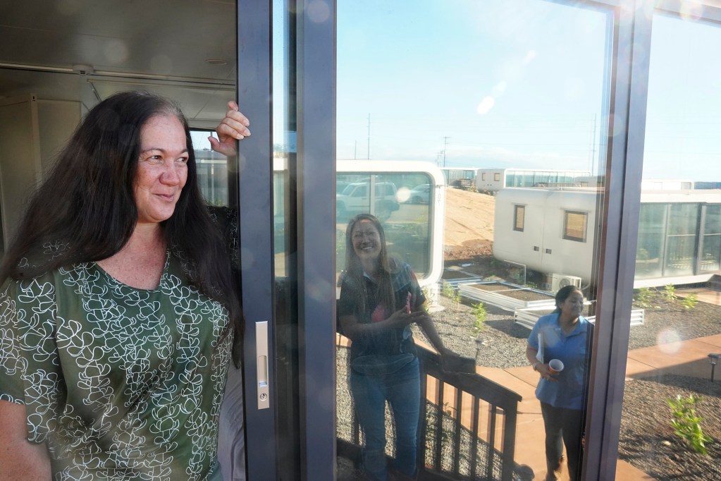 Lana Vierra, from left, smiles when Ka La‘i Ola’s Kalewalani Bancaco mentions Vierra’s daughter lives next door as Joanna Bailey looks on Thursday, Dec. 19, 2024, in Lahaina. Vierra daughter and grandchildren’s home is reflected on the walls of her new, temporary home. (Kevin Fujii/Civil Beat/2024)