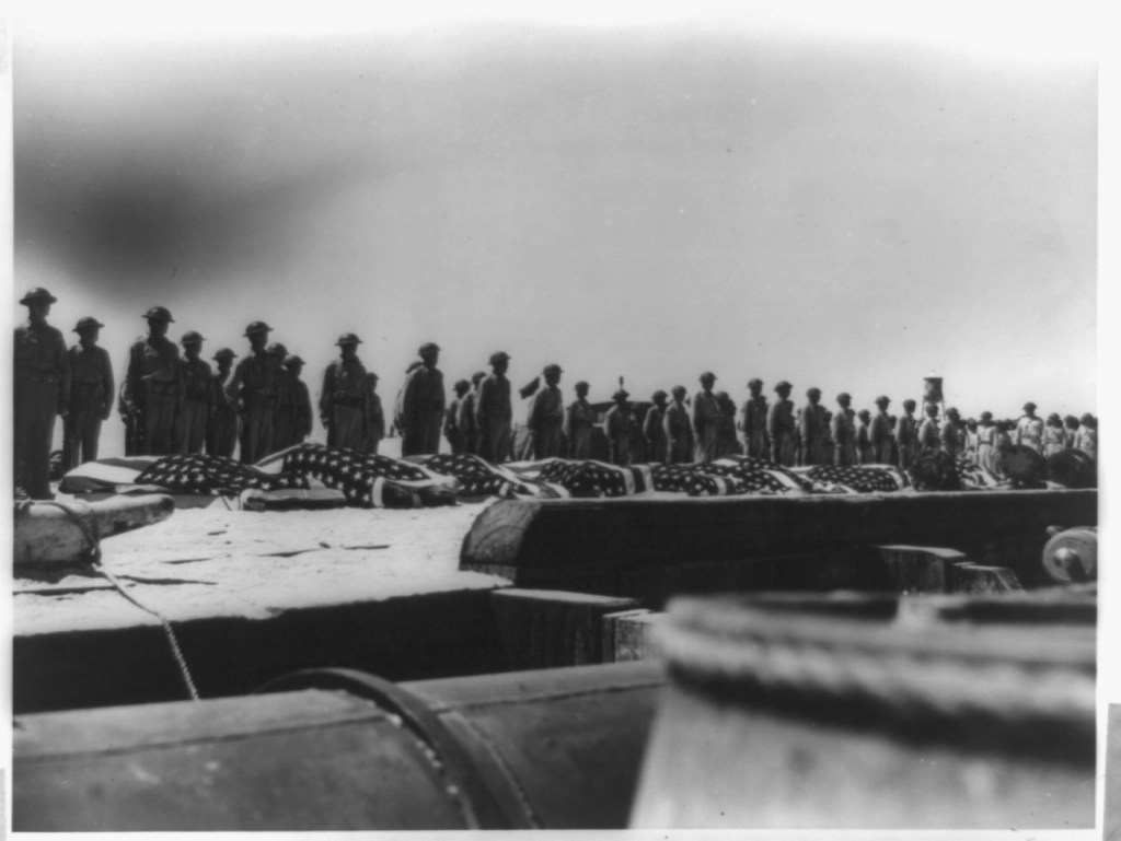 Soldiers standing in a row behind line of flag-draped bodies.