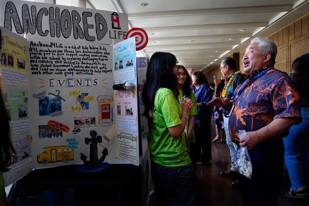 Honouliuli Middle School seventh-graders Isabelle Closa, from left, and Gia Averion surprise Department of Education Superintendent Keith Hayashi with their Education Day display of “Growing Difference Makers in the World Who Know Love, Give Love and Accept Love” at the Capitol Thursday, March 20, 2025, in Honolulu. (Kevin Fujii/Civil Beat/2025)