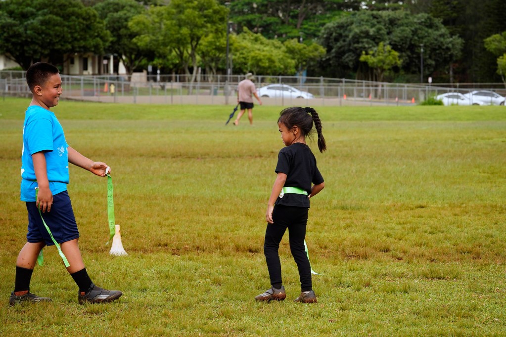 Dawson Arce, 8, gives back a flag to his sister Dezlynn Arce, 5, during flag-football practice at Mililani Mauka Community Park Wednesday, April 16, 2025, in Mililani. Parking vehicles for park users has become an issue. Some parents arrive for weekend games at 4:30 a.m. Opening Mililani Middle School’s parking lot, in the background, could help alleviate parking congestion on the street. (Kevin Fujii/Civil Beat/2025)
