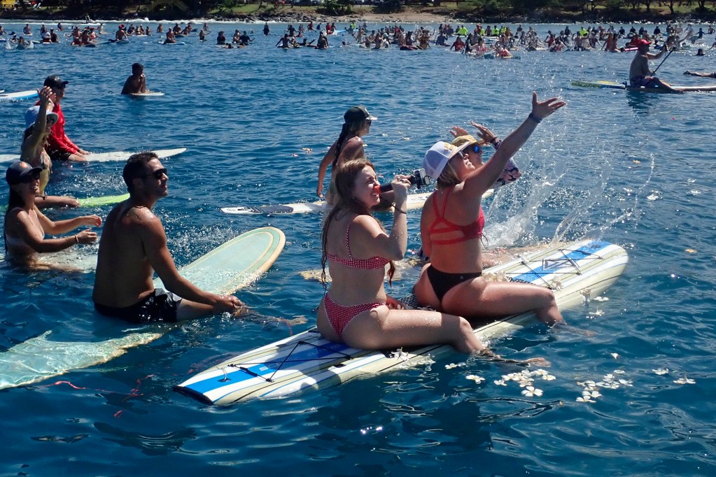 Memorial paddle-out participants from West Maui Kat Lawson films the helicopter dropping flowers while Jennie Hammatt waves off Hanaka‘ō‘ō Beach Park Friday, Aug. 8, 2025, in Lahaina. 102 people died in the Aug. 8, 2023, fire which destroyed the historic West Maui town and Upcountry. (Kevin Fujii/Civil Beat/2025)