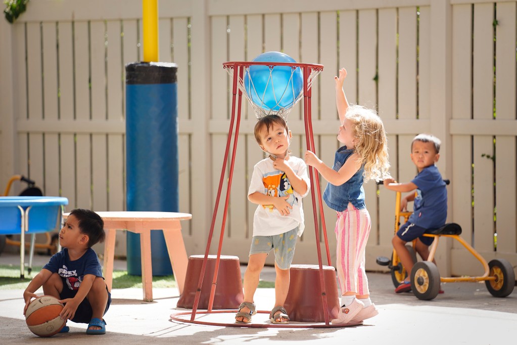 Waikīkī Community Center Preschool students Rian Morrissey, center, stands under the hoop as Zuzu Sheets drops in a ball on the playground Thursday, Sept. 11, 2025, in Honolulu. Julian Rubio, far left, and Aiden Lee, on the tricycle, look on. (Kevin Fujii/Civil Beat/2025)