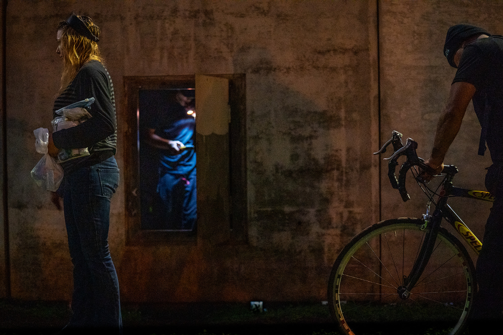 Heather, left, stands outside of a Moanalua Freeway onramp while being interviewed by IHS for the Point In Time homeless count Monday, Jan. 26, 2026, in Honolulu. The person in blue with flashlight inside the access door and the person with the bicycle also stay under the onramp with Heather. (Kevin Fujii/Civil Beat/2026)