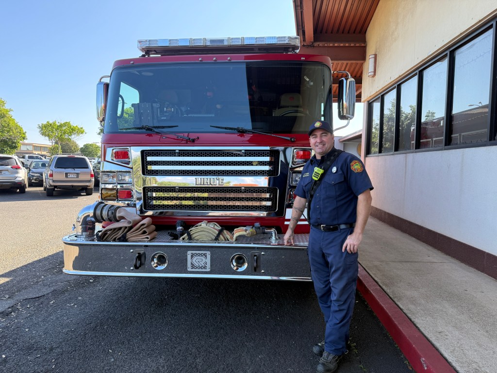 A fire truck from the Lihue fire station is parked outside a building. A firefighter leans on the front of the truck.