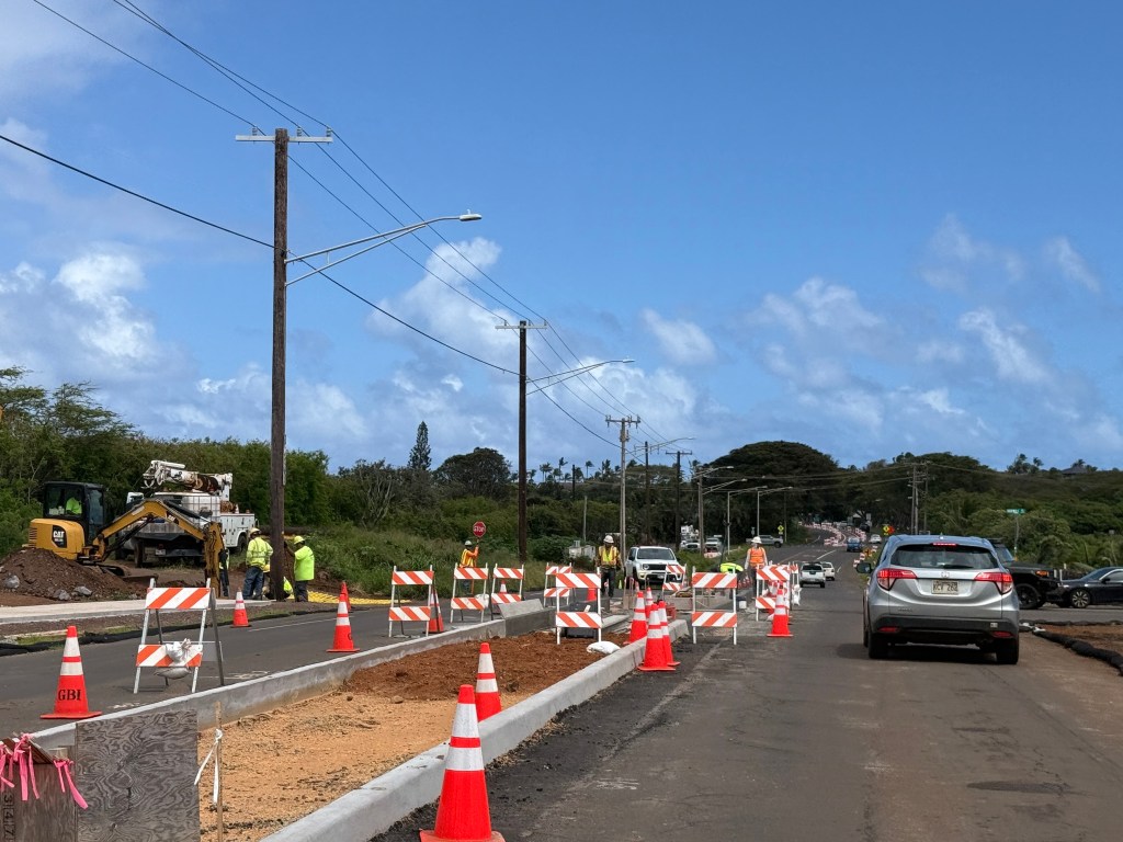 A car waits while orange cones surround a newly constructed cement median in the middle of the road. On the side, workers in bright yellow shirts look toward the ground.