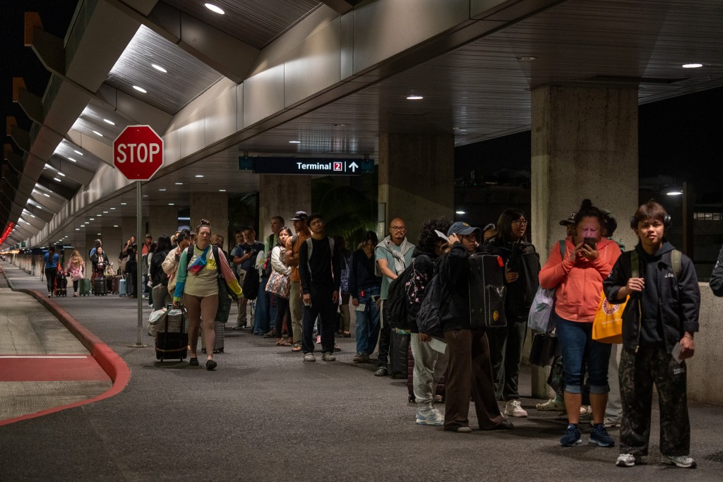 A ragged line of individuals waiting in a line next to concrete pillars to the right and a stop sign to the left. A woman wearing a light, multi-colored long sleeve top walks towards the camera with a roller bag pulled by her right hand.