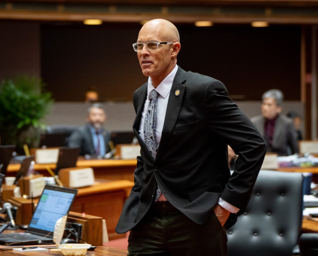 A balding man in a gray suit and light gray tie with his hands in his pockets. He is standing in a room where other men can be seen in the background and a computer is on and open in the lower let.