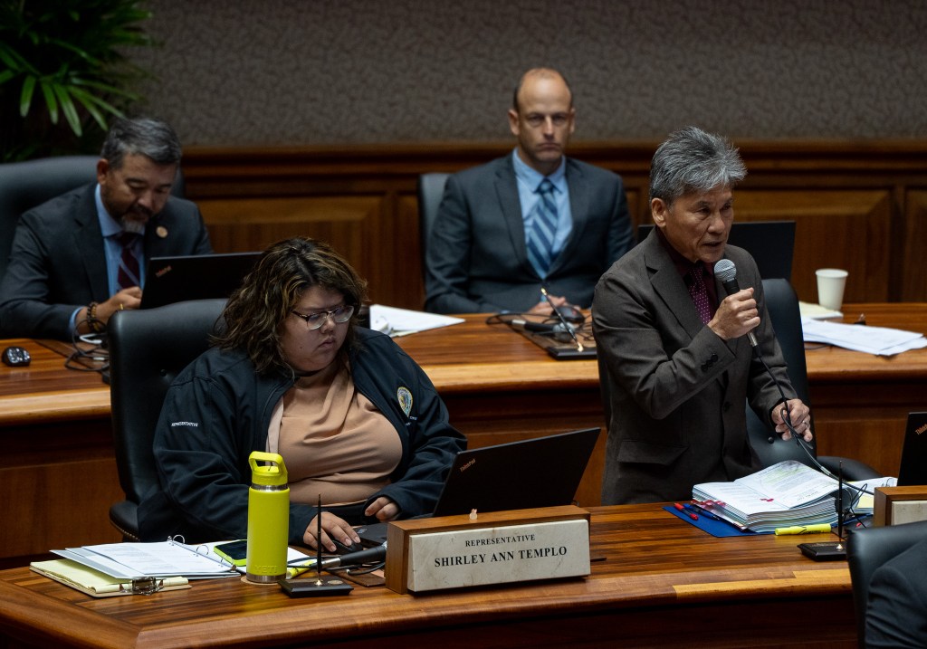 A man with a gray suit and graying hair holds a microphone, right, as other individuals sit around him. A woman sits to the left and two men are seated wearing jackets and ties in the background.