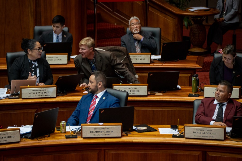 Seven individuals seated at three rows of desks, with the man in the center of the photo speaking to a woman at left.