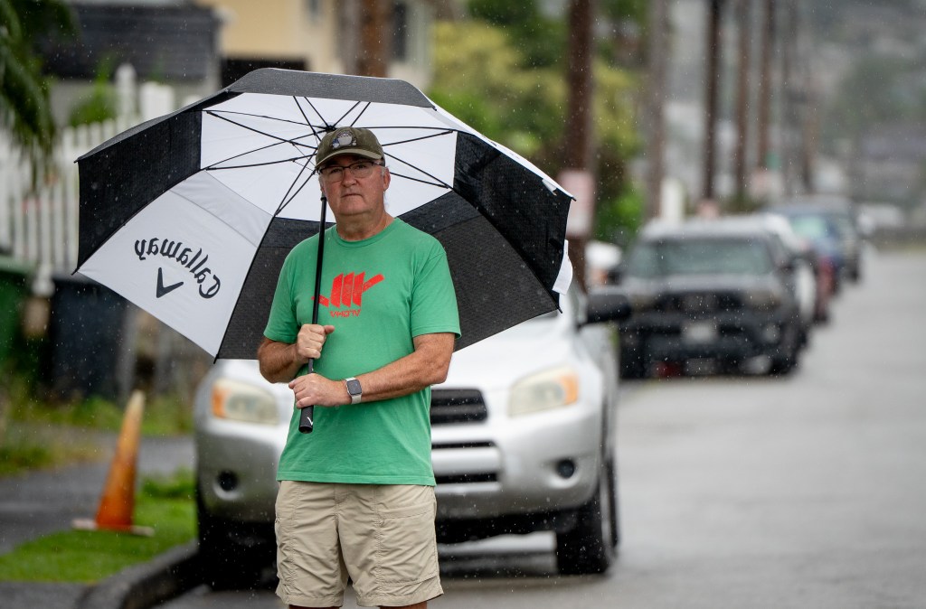 A man wearing a green t-shirt stands with a large black and white umbrella on a street with cars out of focus in the background.