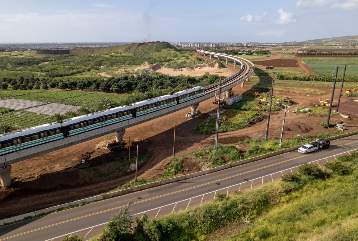 An elevated rail line and train are seen next to a roadway surrounded by farm and other less developed land.
