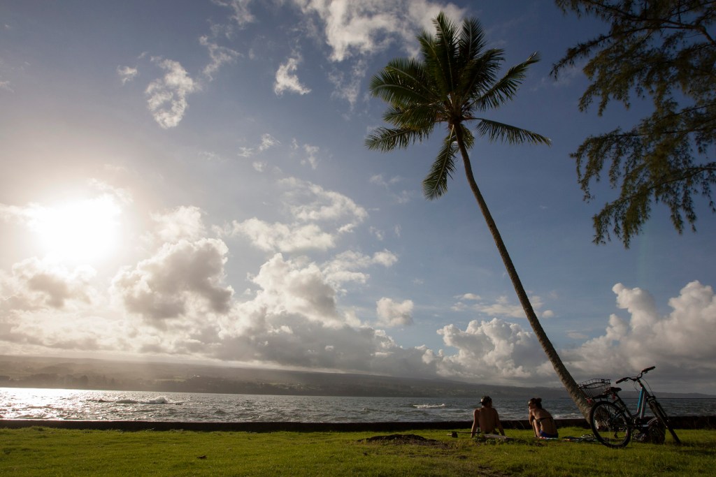 FILE - In this Aug. 30, 2015, file photo, Keenin Ide, left, of Hilo, Hawaii, and Medea Yankova, of Sofia, Bulgaria, sit near Hilo Bay in Hilo, Hawaii. The Big Island visitors bureau and Hawaii County have launched a campaign that aims to create a more conscientious tourism industry on the Big Island. The Island of Hawaii Visitors Bureau Executive Director Ross Birch and county Managing Director Will Okabe presented the new "Pono Pledge" at a Thursday, Sept. 20, 2018, event, the Hawaii Tribune-Herald reported.  (AP Photo/Caleb Jones, File)