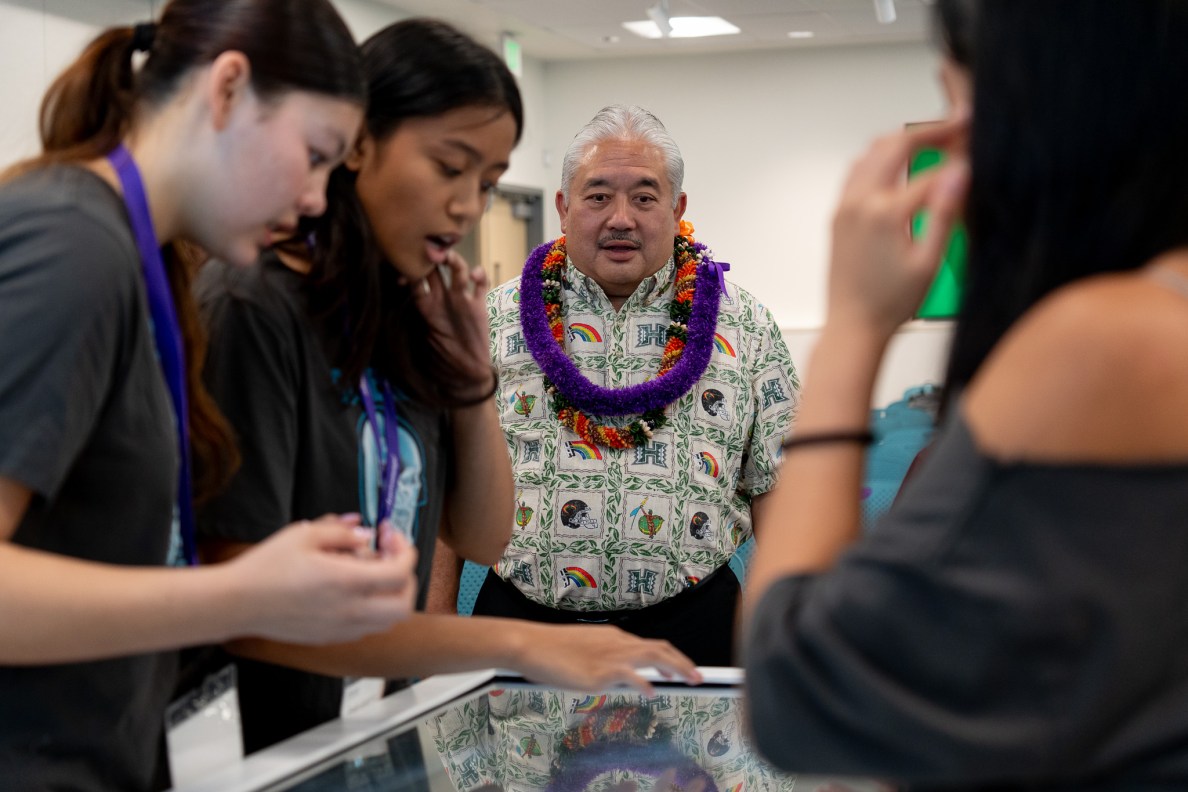 A man wearing a Hawaiian shirt and lei watches students wearing gray t-shirts work on a table top device.