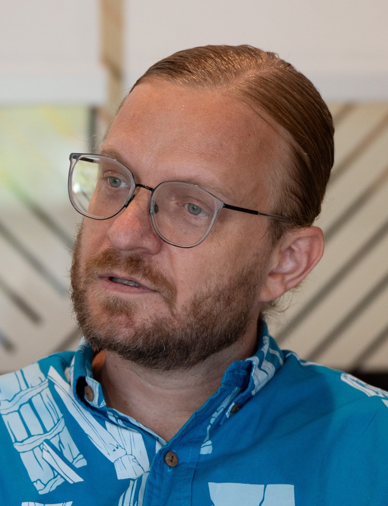 A light-skinned man with light brown hair tightly pulled back. He has a beard, wears glasses and a blue patterned shirt.