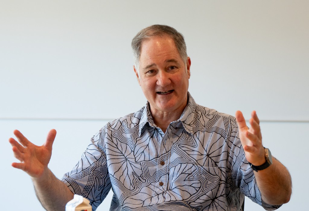A light-skinned man gestures while speaking. He wears a patterned gray shirt and wears a watch on his left wrist.