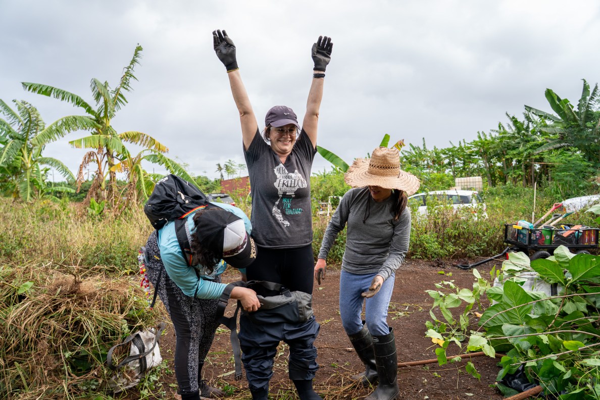 Megan Dooley gets a hand putting on waders during work clearing a ditch of overgrown trees and foliage at Mill 6 Farms in Waialua May 23, 2026. Rebecca Stringer, left, and Julie Taniyama lend a hand. (Craig Fujii/Civil Beat/2026)