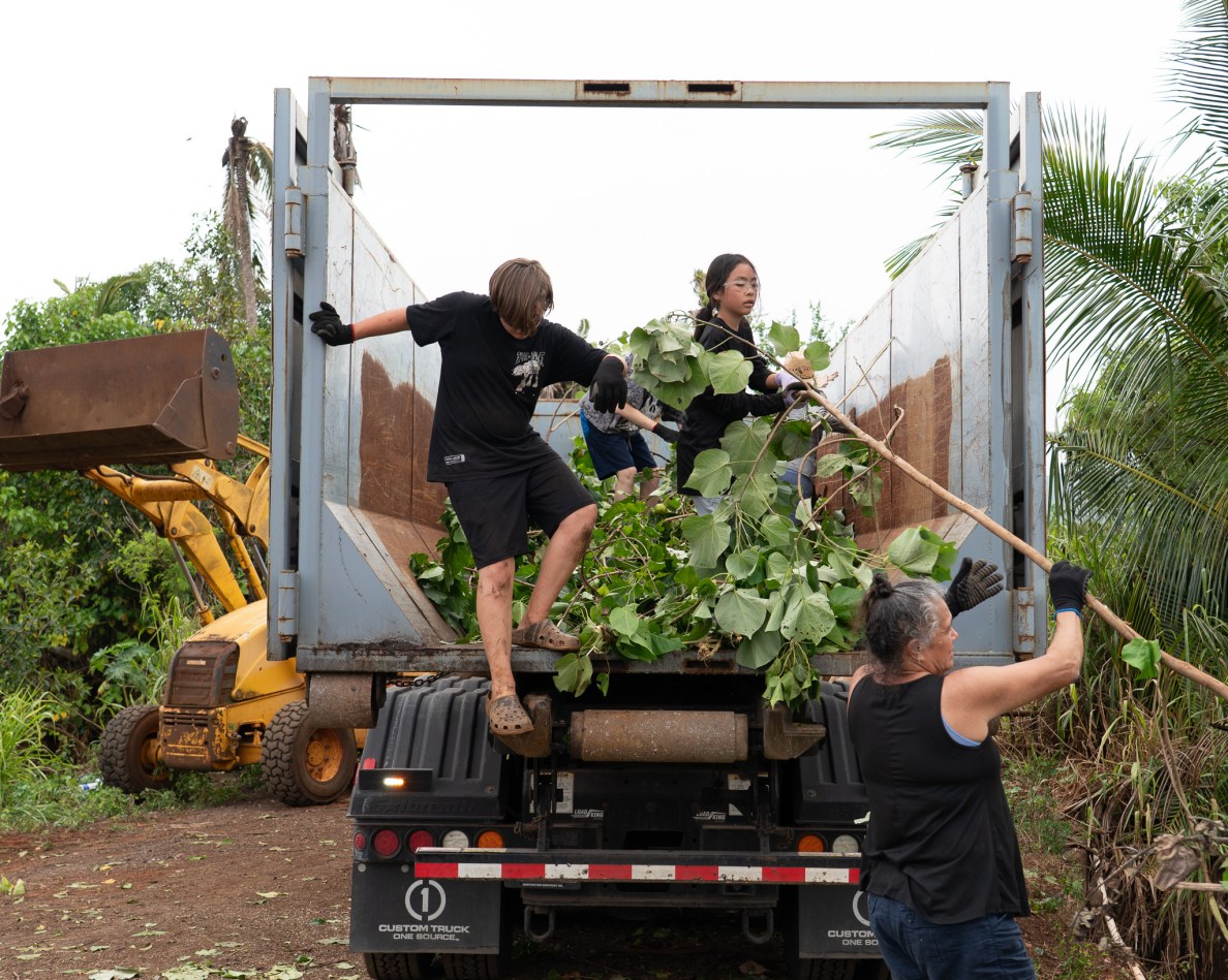 Markus Searls, left, steps down from a roll-off dumpster as he works alongside Lauren Taniyama at Mill 6 Farms in Waialua May 23, 2026. The two worked with volunteers to clear ditches of overgrown trees and plants to improve water flow after the Kona low storms flooded the area. (Craig Fujii/Civil Beat/2026)