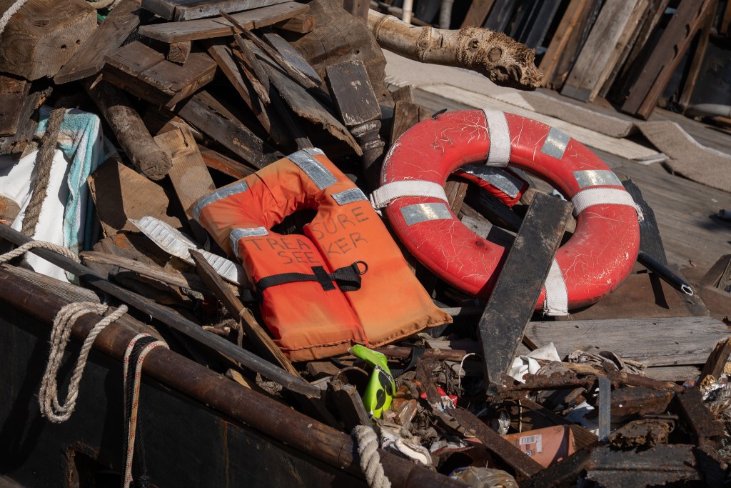 An orange life jacket and an orange flotation ring in the middle of a pile of broken timber steel and other rubbish.